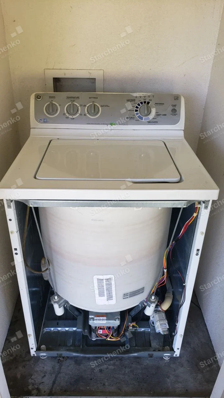 GE logo at upper left of washer control panel, front removed exposing tub and lower drive components.