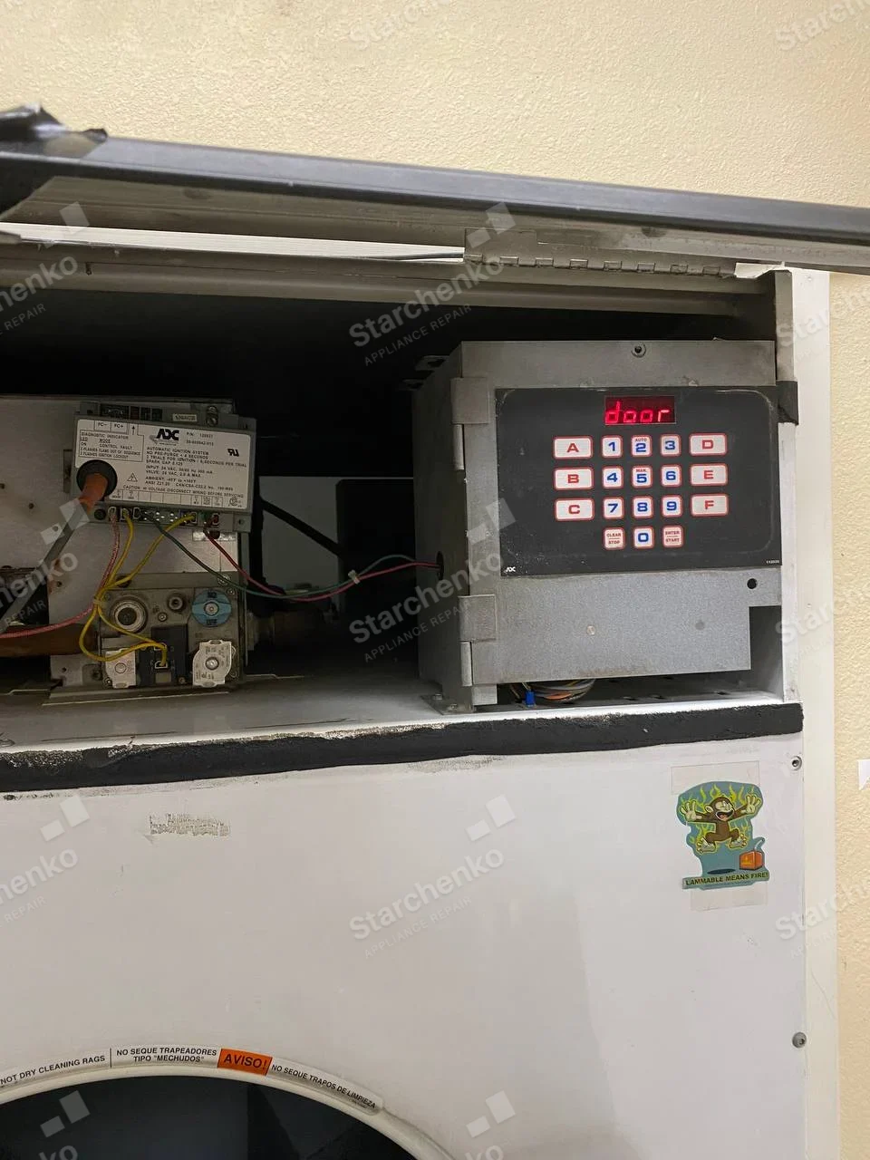 American Dryer (ADC) repair: Upper service door of large dryer open, exposing gas valve and electronic keypad/control, warning near drum opening says not to dry cleaning rags.