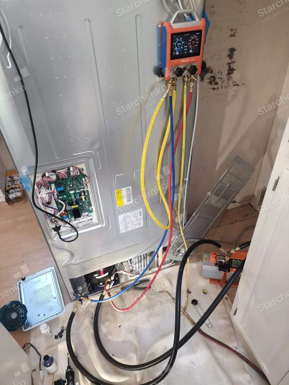 KitchenAid repair: Rear of refrigerator with exposed control board and compressor, connected service gauges and hoses.