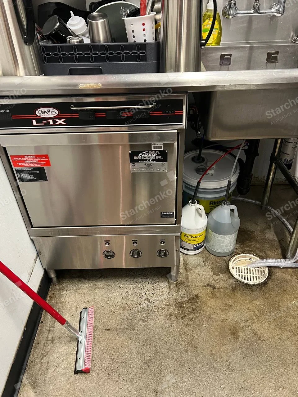 CMA logo and L-1X label on undercounter dishwasher front control fascia, chemical jugs next to it.