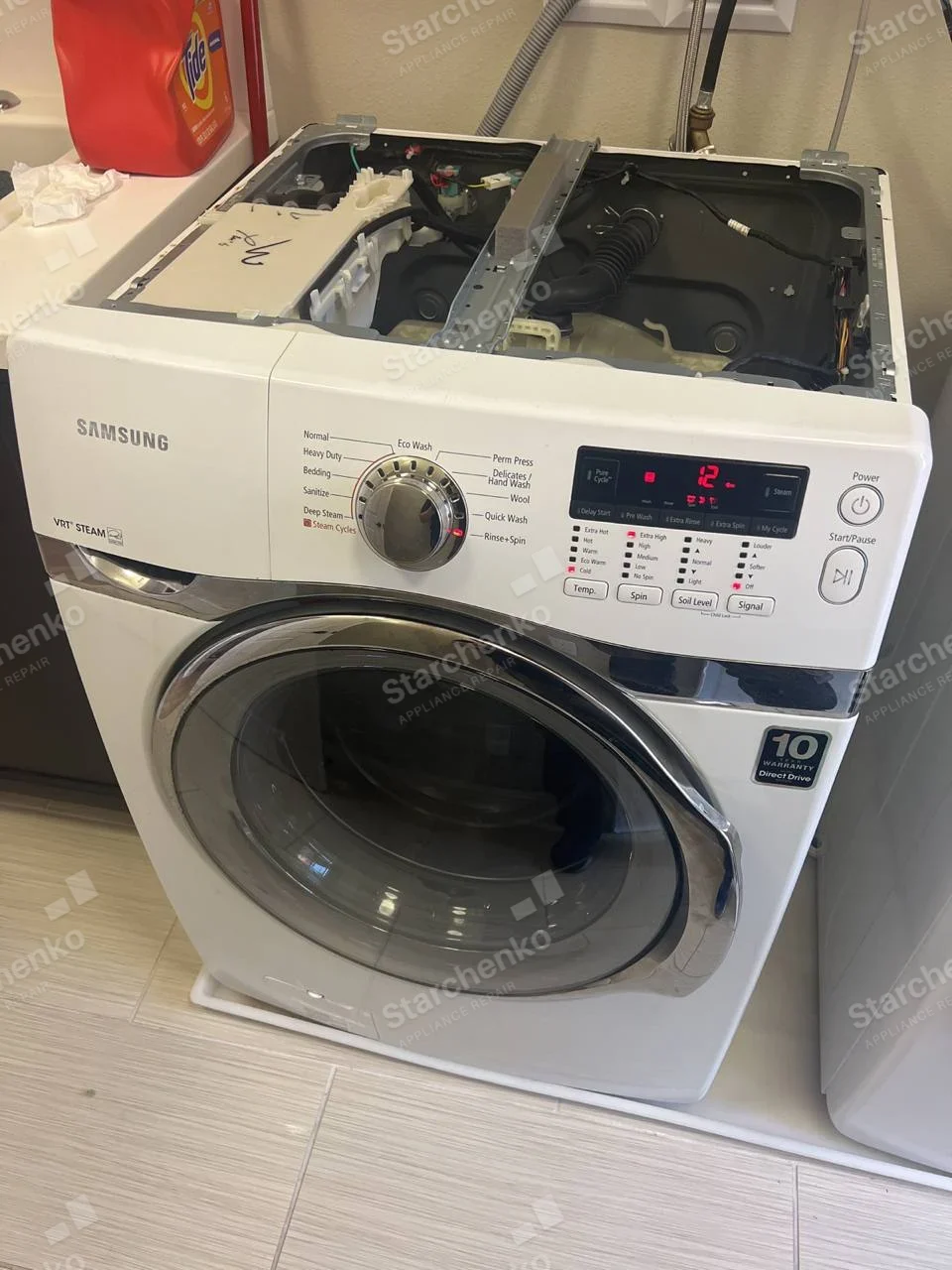 Samsung front-load washer with top cover removed, exposing tub, dispenser and hoses.