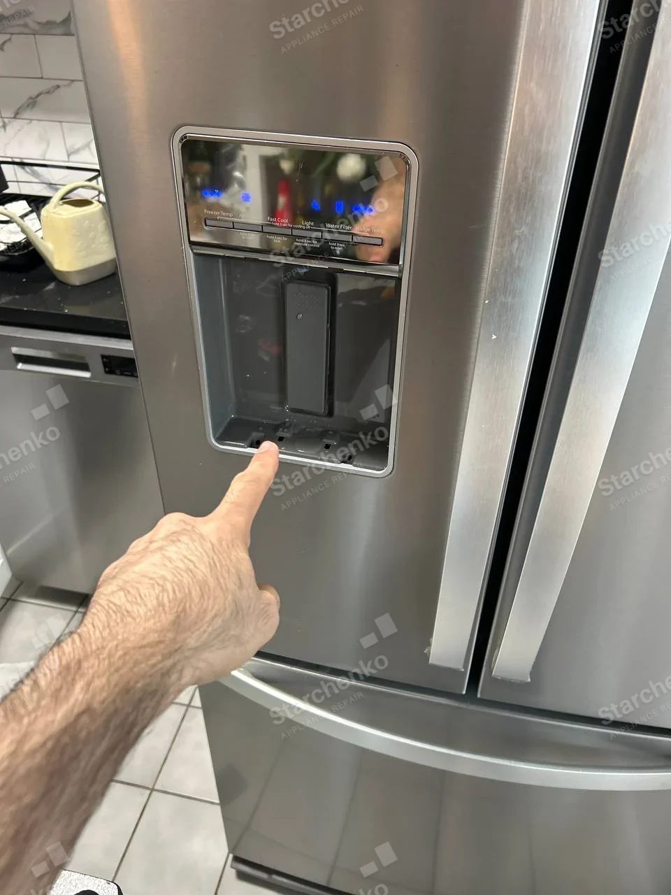 KitchenAid repair: French-door refrigerator with dispenser trim removed and technician pointing at the dispenser recess.