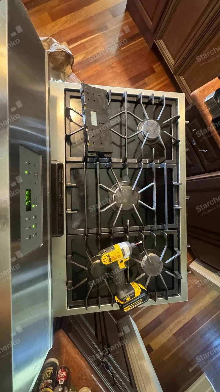 Amana repair: Top of gas range with burner assembly and cooktop grates visible, drill placed on surface, no legible main-appliance badge.