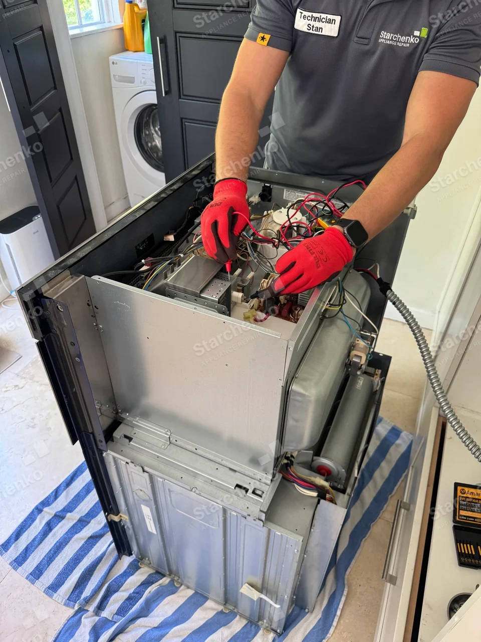 Electrolux repair: Technician probes wiring in the opened back of a cooking appliance with door glass and insulation, LG washer and Amana package are adjacent, not the repair target.