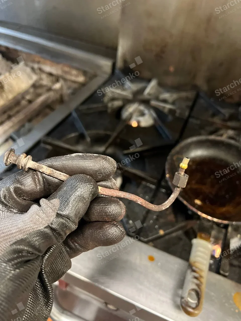 Southbend repair: Technician holds bent gas pilot/thermocouple tube in front of heavy cast-iron burners and a lit flame.