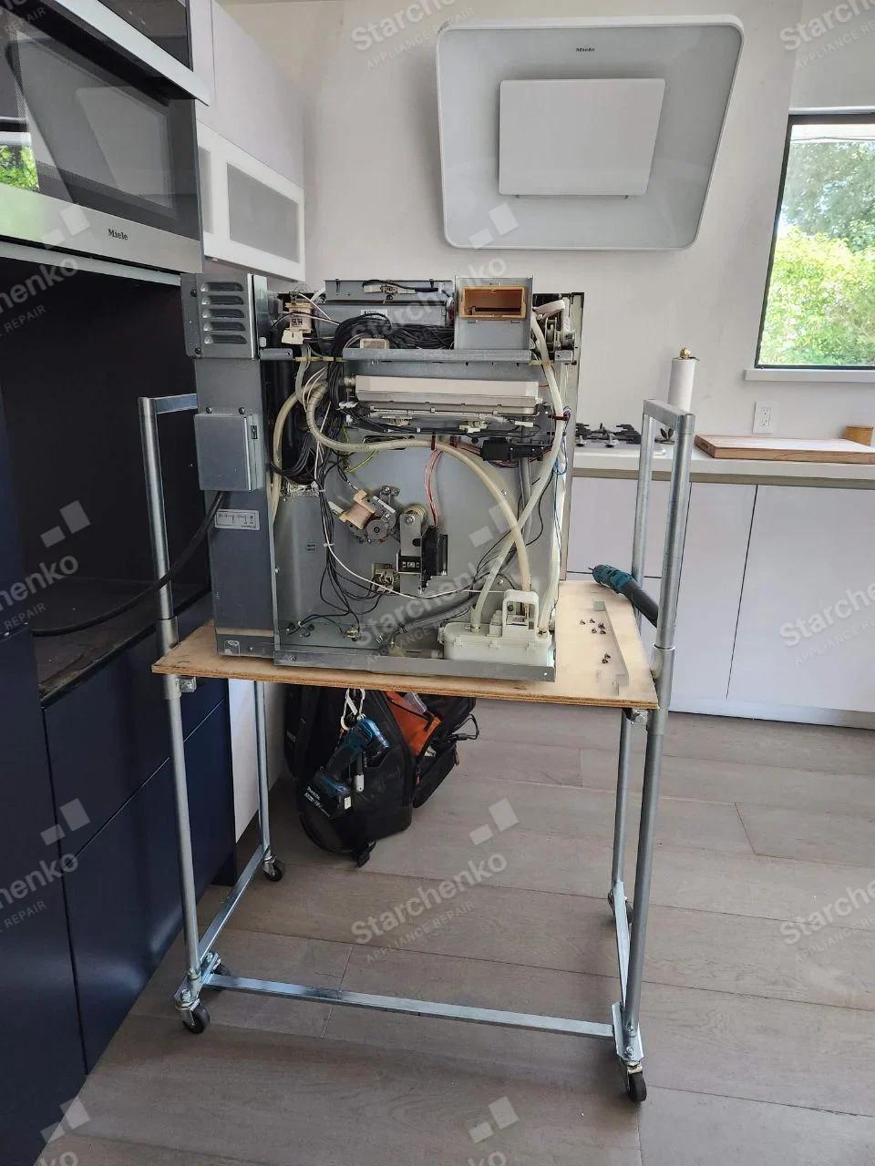 LG repair: Dishwasher body on service cart with rear shell removed exposing hoses, wiring and pump, Miele lettering belongs to adjacent microwave and range hood only.