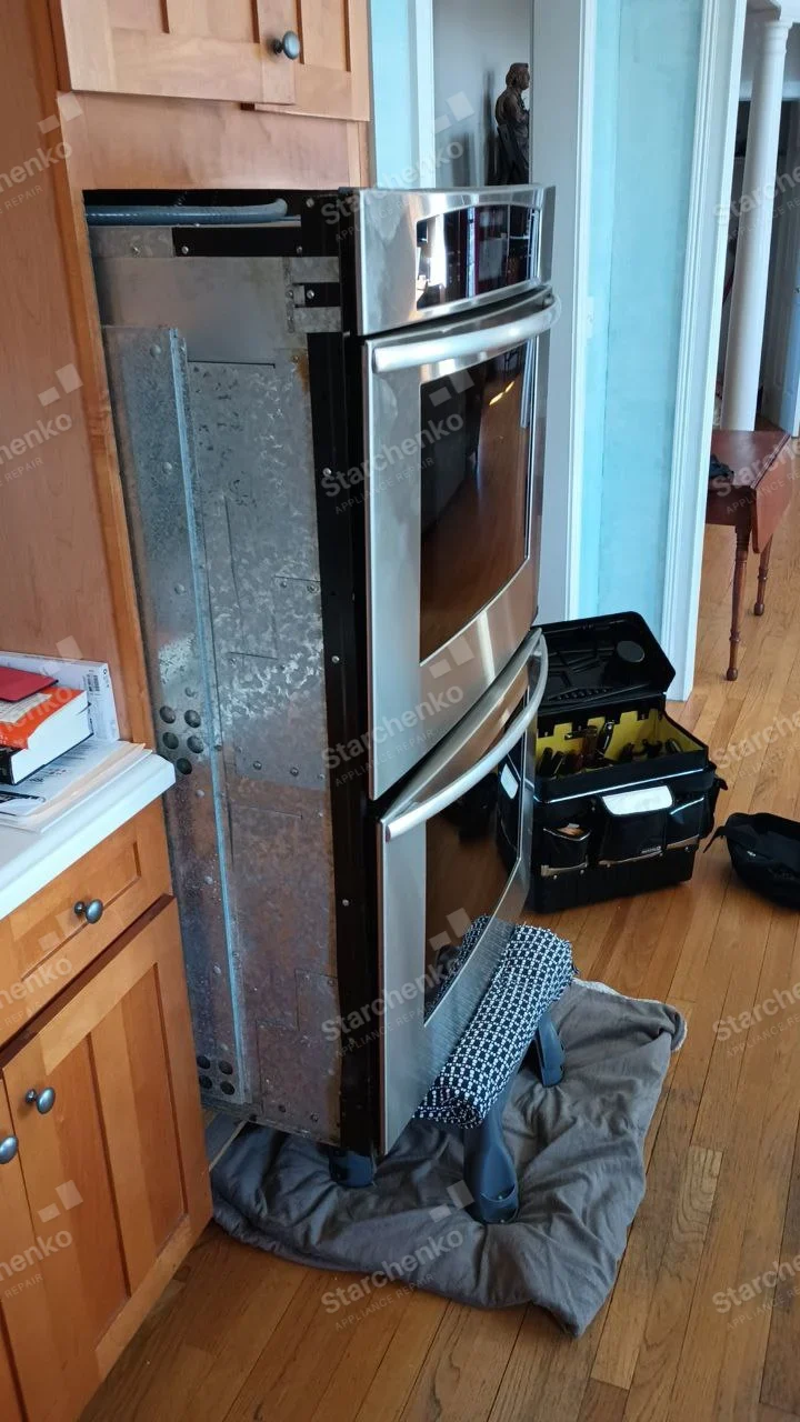 GE repair: Stainless double wall oven pulled from cabinet with left metal side exposed on protective blanket.