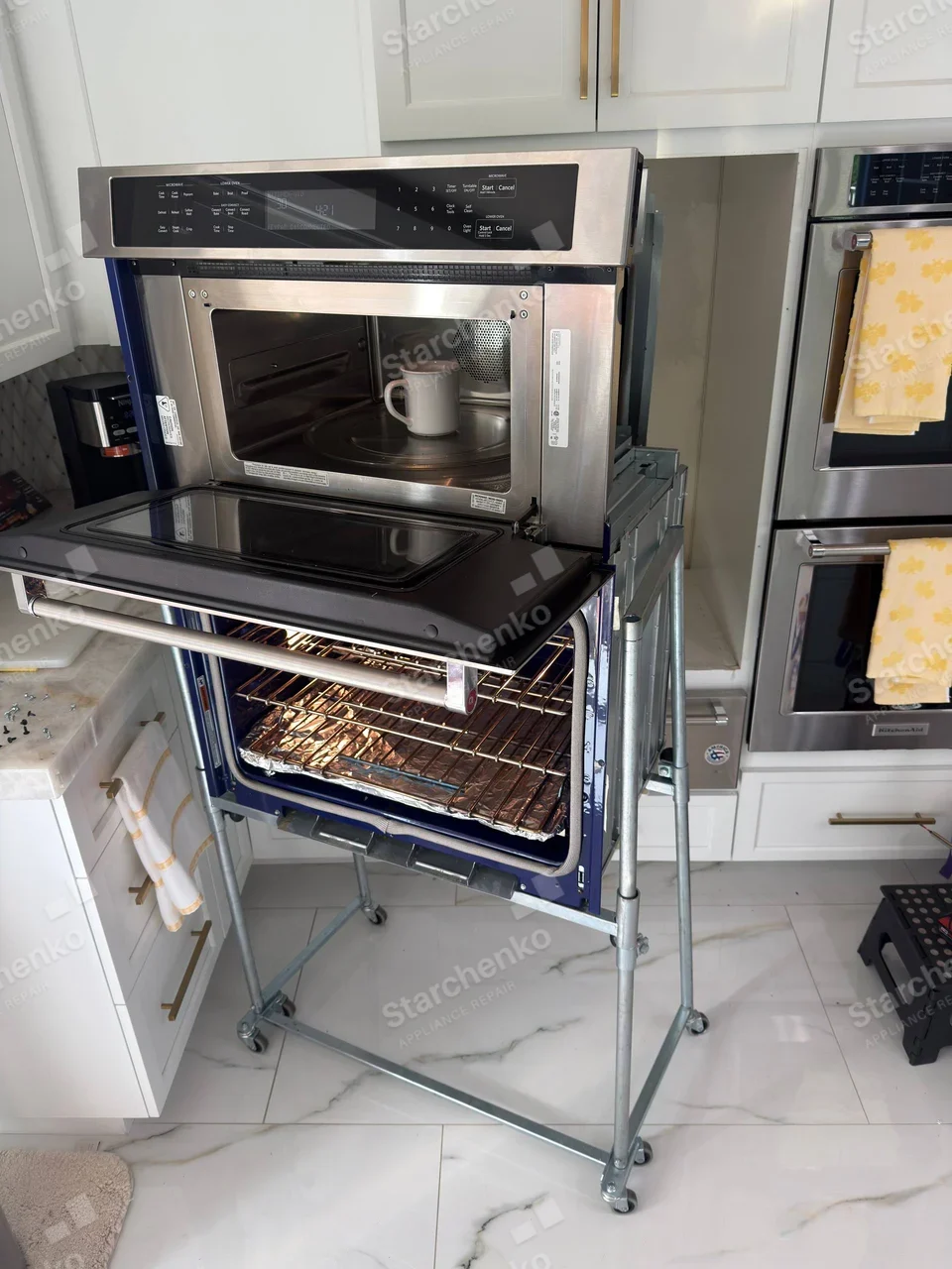 Pulled-out microwave above open wall oven, neighbouring double oven has KitchenAid logo, but the pulled-out unit's own brand is not independently legible.