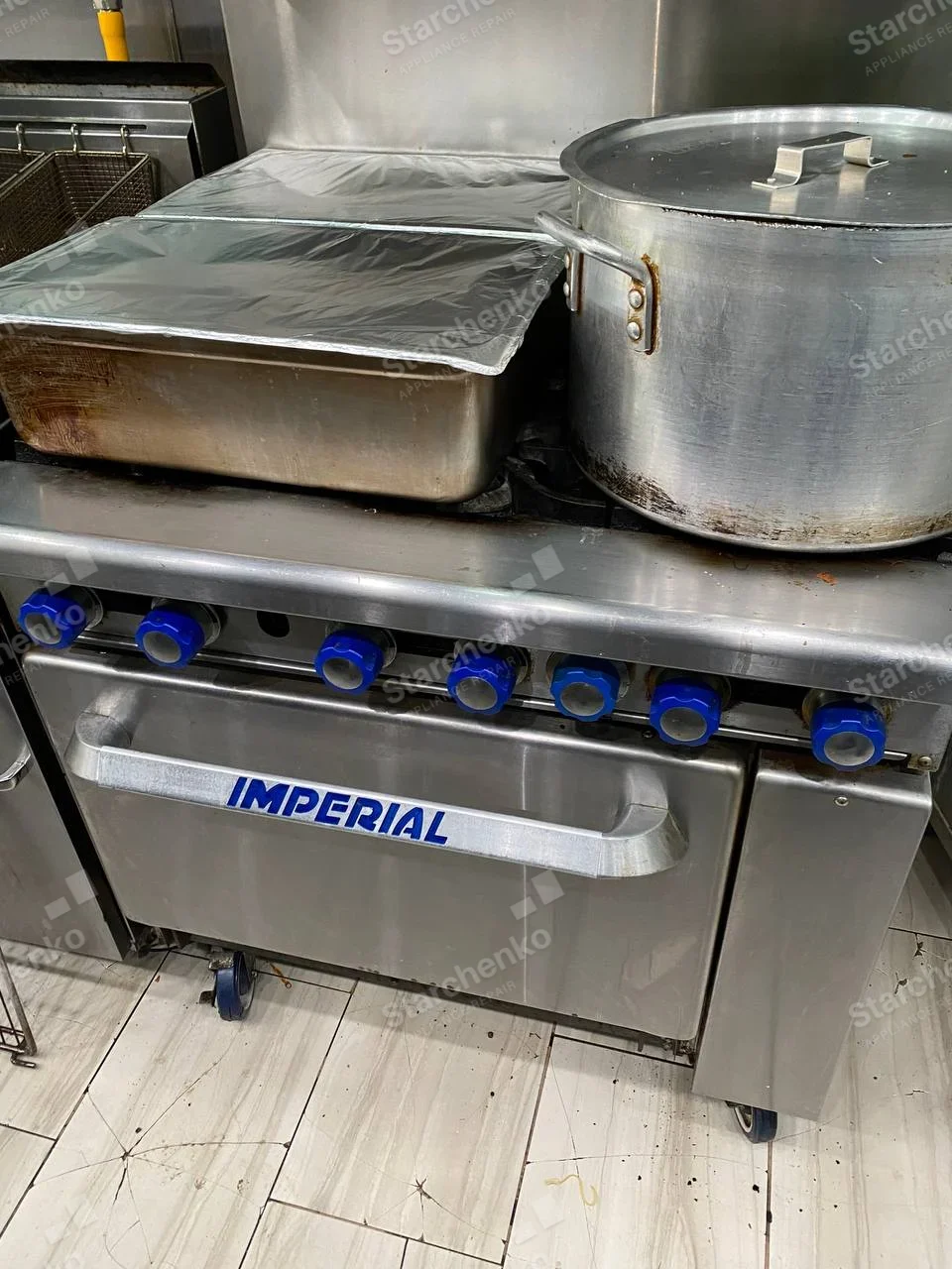 IMPERIAL lettering across oven door handle of heavy stainless commercial range with row of blue burner knobs.
