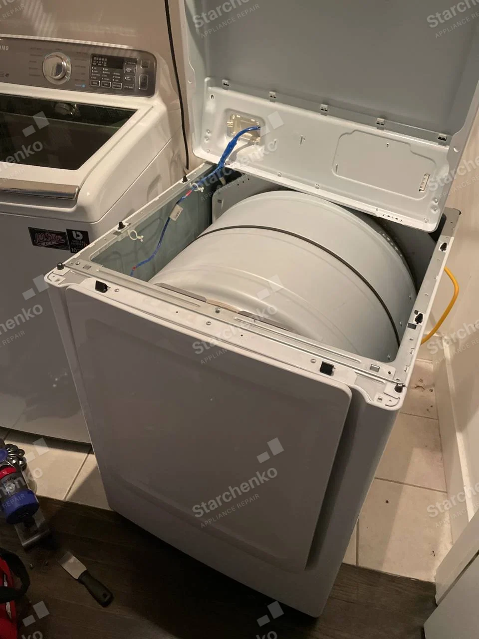 White Samsung dryer with top and side panels removed, exposing large drum shell beside a washer.