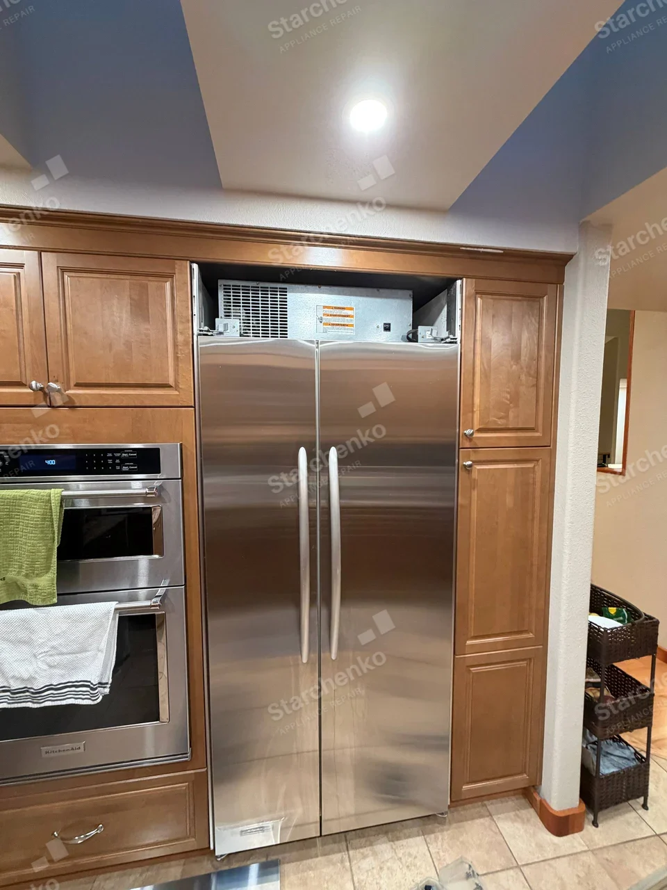 Built-in GE Profile side-by-side refrigerator with upper machine compartment exposed, KitchenAid badge belongs to separate adjacent oven.