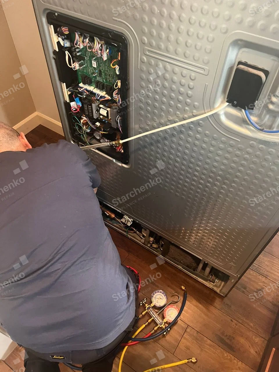 Maytag repair: Technician accesses electronic board and refrigeration lines behind large textured rear panel, manifold gauges on floor.