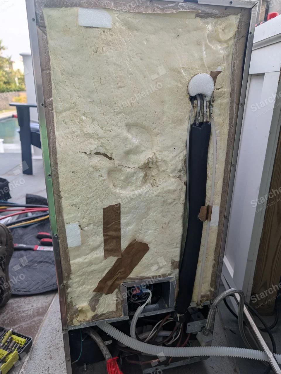 Undercounter ice maker pulled out with its rear panel removed, showing foam insulation and water lines