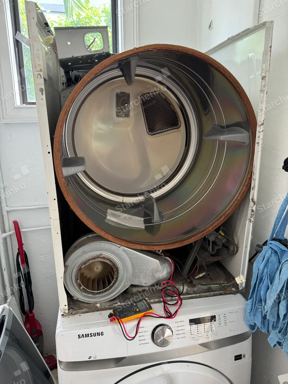 Stacked dryer with the drum pulled out and multimeter leads connected inside the cabinet, above a front-load washer